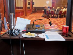 Afgebeeld staat een vertaalhok, met op tafel een microfoon, een koptelefoon, papieren en een bord met koekjes. Het Nederlands Genootschap van Tolken en Vertalers (NGTV) bestaat 65 jaar. Deze mijlpaal is voor de vereniging en haar 1000 leden een stimulans om aandacht te blijven vragen voor het professionele vak van vertalers en tolken. Daarom vieren zij het jubileum groots met een reeks boeiende (online) activiteiten, waaronder de workshops ‘Loesje’ en ‘Muziek en Taal’ met bekende zanger Jan Rot en tolk Nederlands Gebarentaal Mirjam Stolk.