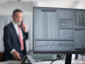 2021 eindigde met een 28% hogere AEX dan in 2020 en was zo het beste investeringsjaar ooit voor beleggers in aandelen. | Afgebeeld staat een desktopscherm met statistieken. Op de achtergrond staat geblurd een witte man met donker haar en een grijze baard gekleed in een donkerblauw pak, wit overhemd en een lichtroze stropdas. Hij telefoneert. | pexels kampus production 8353775