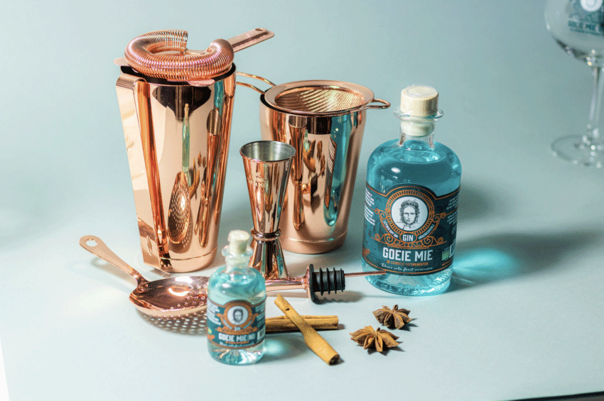 Goeie Mie Gin haalt investering op bij Dragons’ Den - afgebeeld staan twee flesjes Goeie Mie Gin omgeven door cocktailshakers en andere cocktailmaterialen, sterrenmunt en kaneelstokjes. 