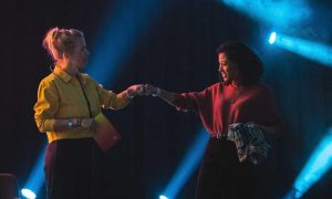 Twee vrouwen pakken elkaars hand. Ze staan voor een zwarte achtergrond met blauwe lichten. Techneuten en ondernemers werken aan de toekomst van het internet, maar consumenten tasten in het duister. Wat heeft de toekomst in petto? Verschillende experts die spreken tijdens The Next Web Conference in Amsterdam op 16 & 17 juni, delen hun verwachtingen van de toekomst van het internet.